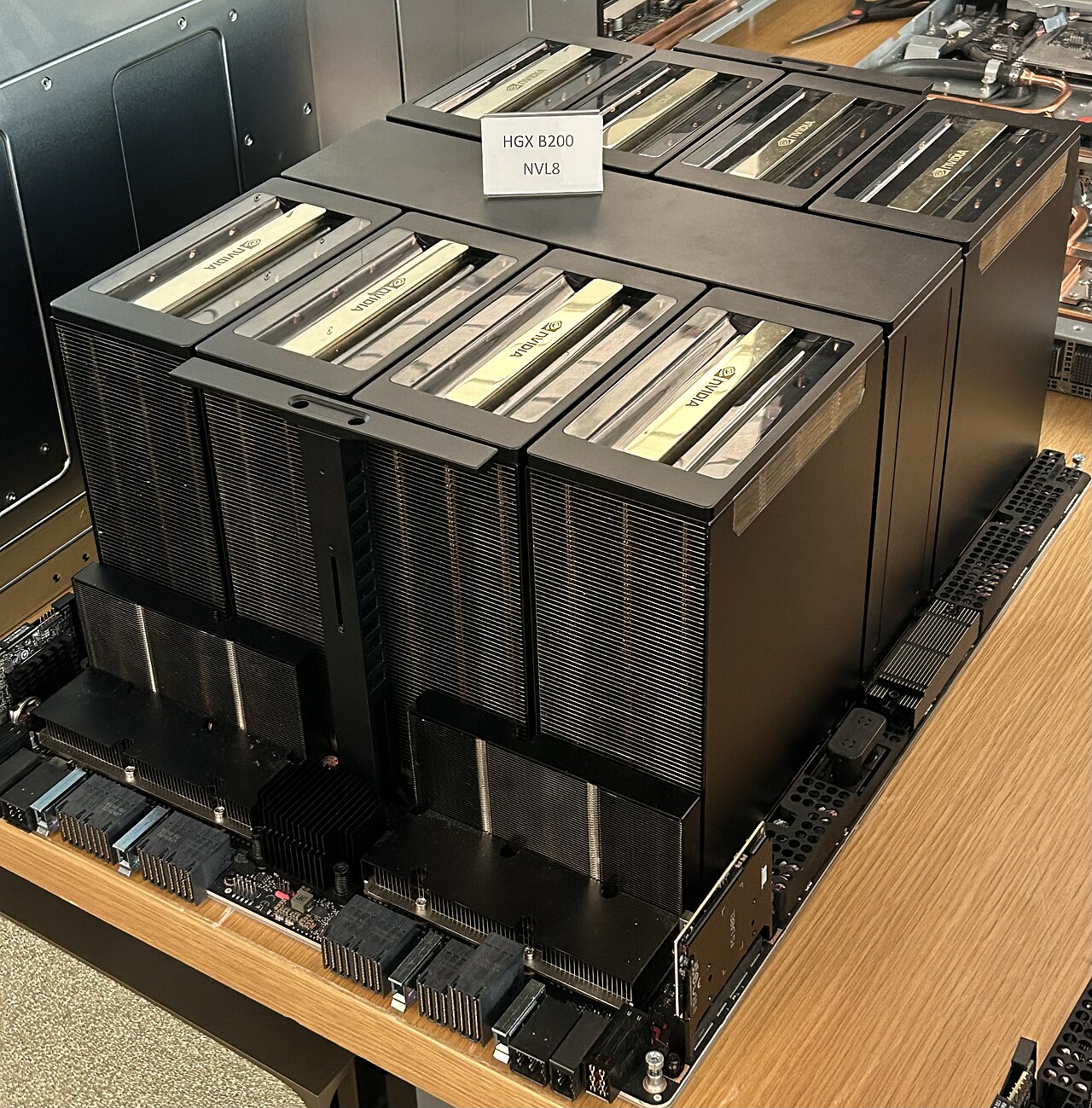 Photograph of an HGX B200 baseboard on a table: eight tall black GPU heatsinks in two rows of four, lower heatsinks along the front edge, and connectors along the edges